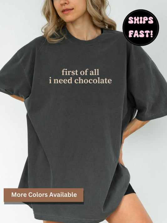 A woman wearing a comfort colors t-shirt in pepper with the text "first of all I need chocolate" printed on it.