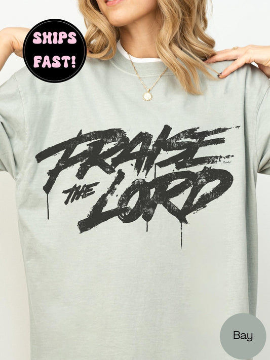 A woman wearing a gray t-shirt with the text "PRAISE THE LORD" printed on it.