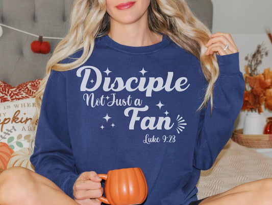 A woman wearing a blue sweatshirt with the text "Disciples Not Just Fans" and a Bible verse reference, "Luke 9:23", is sitting on a bed with a pumpkin-themed pillow and holding a mug.