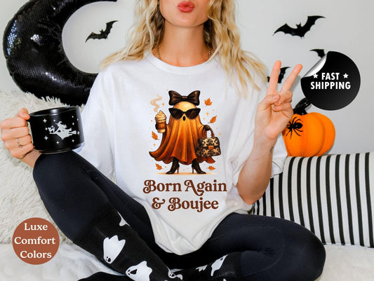 A woman wearing a white t-shirt with a graphic of a witch and the text "Born Again & Boujeee" is sitting on a couch with a mug and a pumpkin decoration.