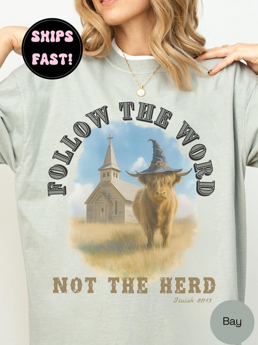 A woman wearing a bay green t-shirt with a graphic design featuring a cow wearing a witch's hat and a church in the background.