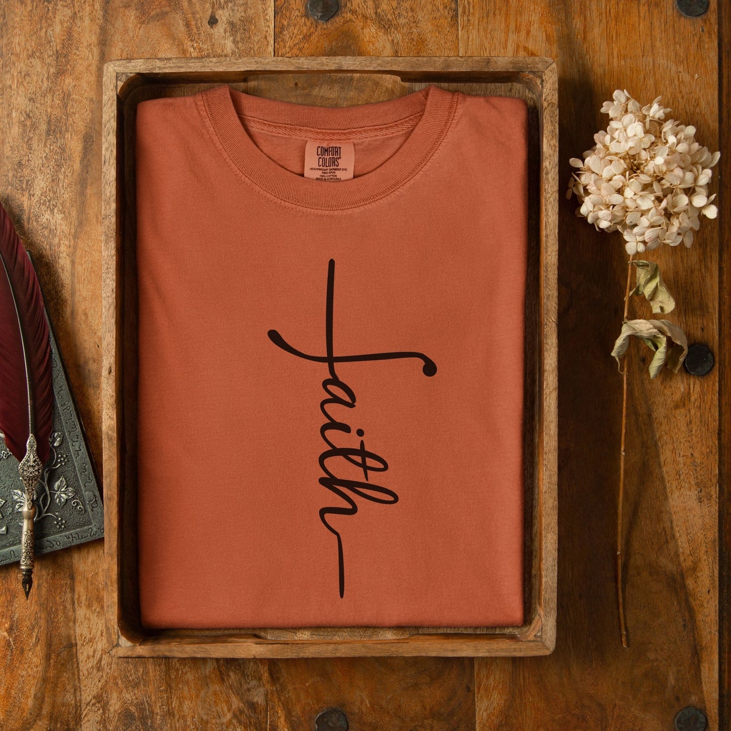 a red t-shirt with the word "Faith" written on it, placed in a wooden box on a wooden surface.
