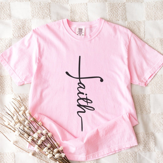 A pink t-shirt with the word "Faith" printed on it, along with a small floral arrangement.