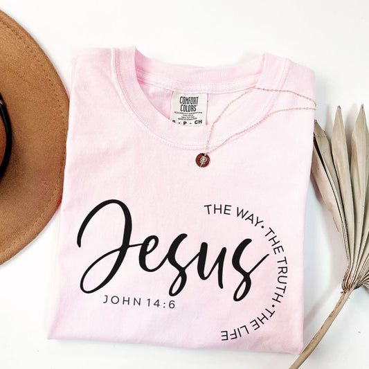 A pink t-shirt with the text "The way. The truth. Jesus" printed on it, along with the Bible verse "John 14:6". The t-shirt is folded and placed next to a brown hat and some dried leaves.