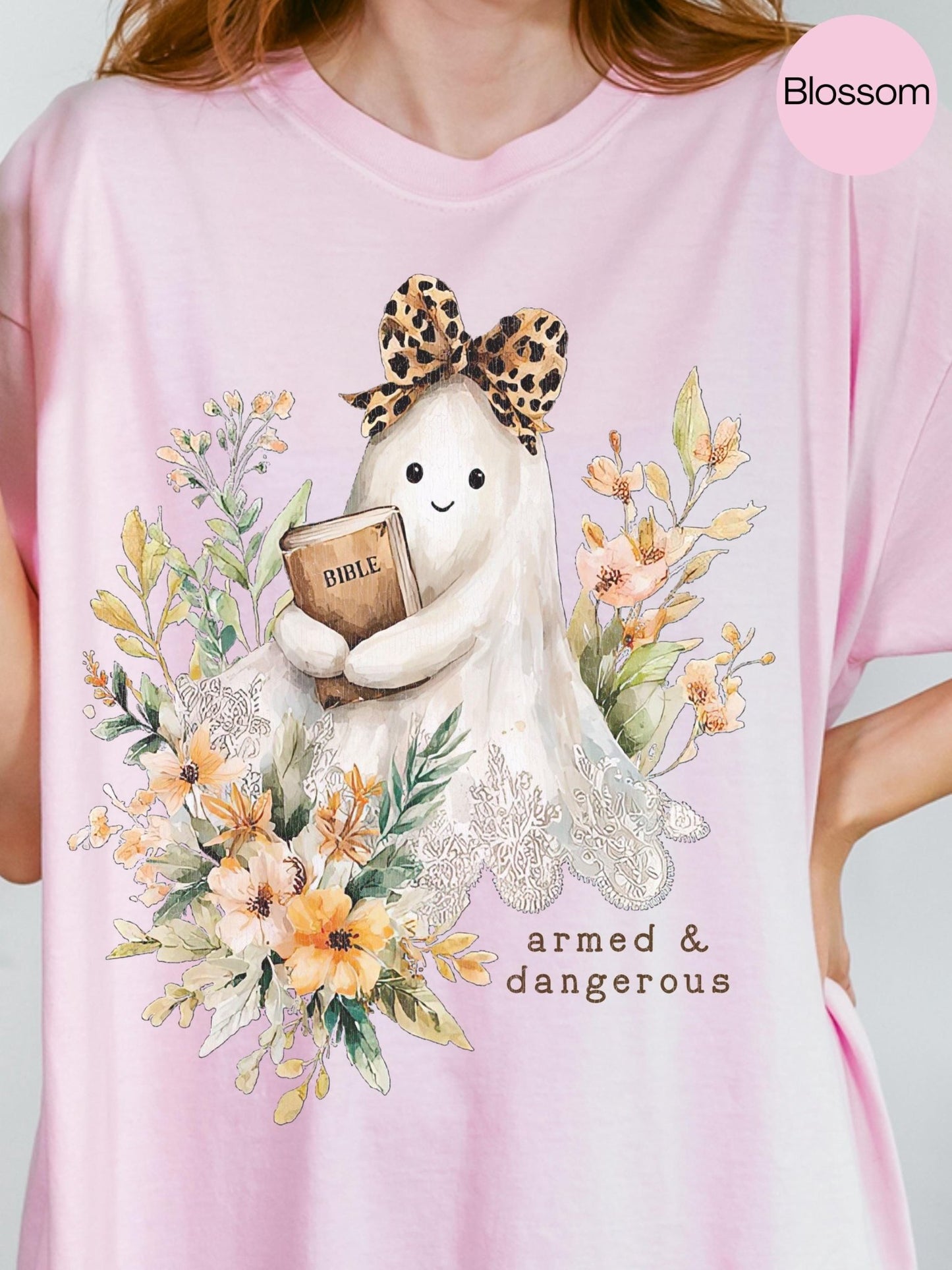 A pink t-shirt with a graphic design featuring a ghost-like figure holding a Bible and surrounded by flowers and foliage.