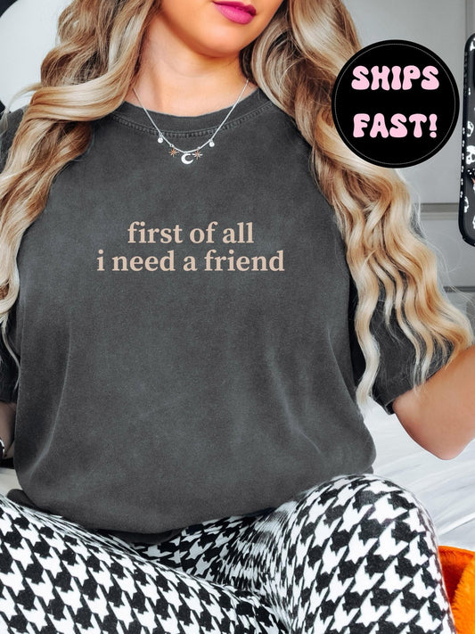 A woman with long, wavy blonde hair wearing a gray t-shirt with the text "first of all I need a friend" printed on it.