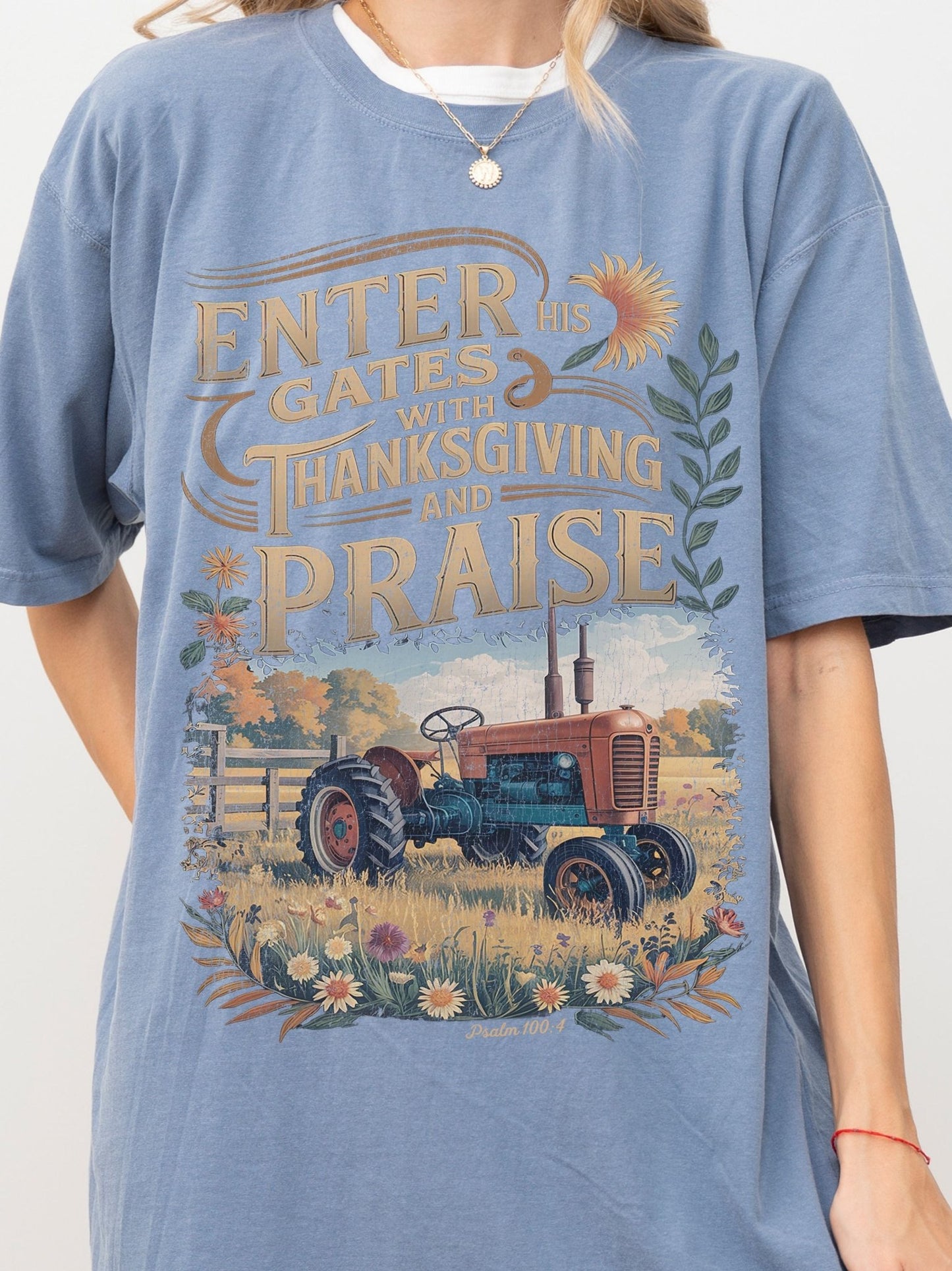 A blue t-shirt with a graphic design depicting a tractor in a field with flowers and a sunflower. The text on the t-shirt reads "ENTER his GATES with THANKSGIVING and PRAISE".