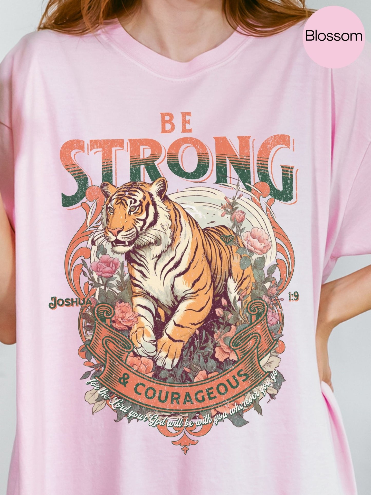 A pink t-shirt with a graphic design featuring a tiger surrounded by flowers and foliage, along with the text "Be STRONG & COURAGEOUS" and a Bible verse reference.
