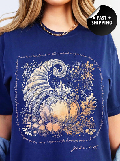 A woman wearing a blue t-shirt with a graphic design depicting a golden cornucopia surrounded by autumn leaves and pumpkins.