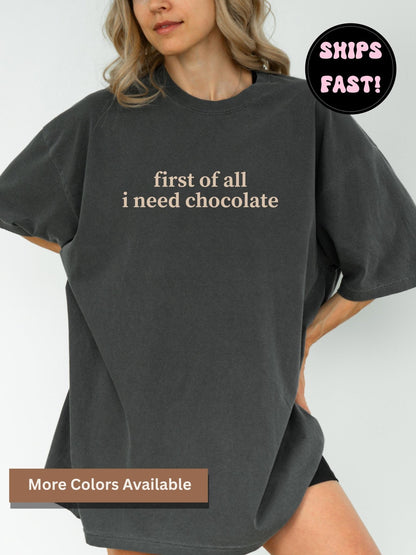 A woman wearing a comfort colors t-shirt in pepper with the text "first of all I need chocolate" printed on it.