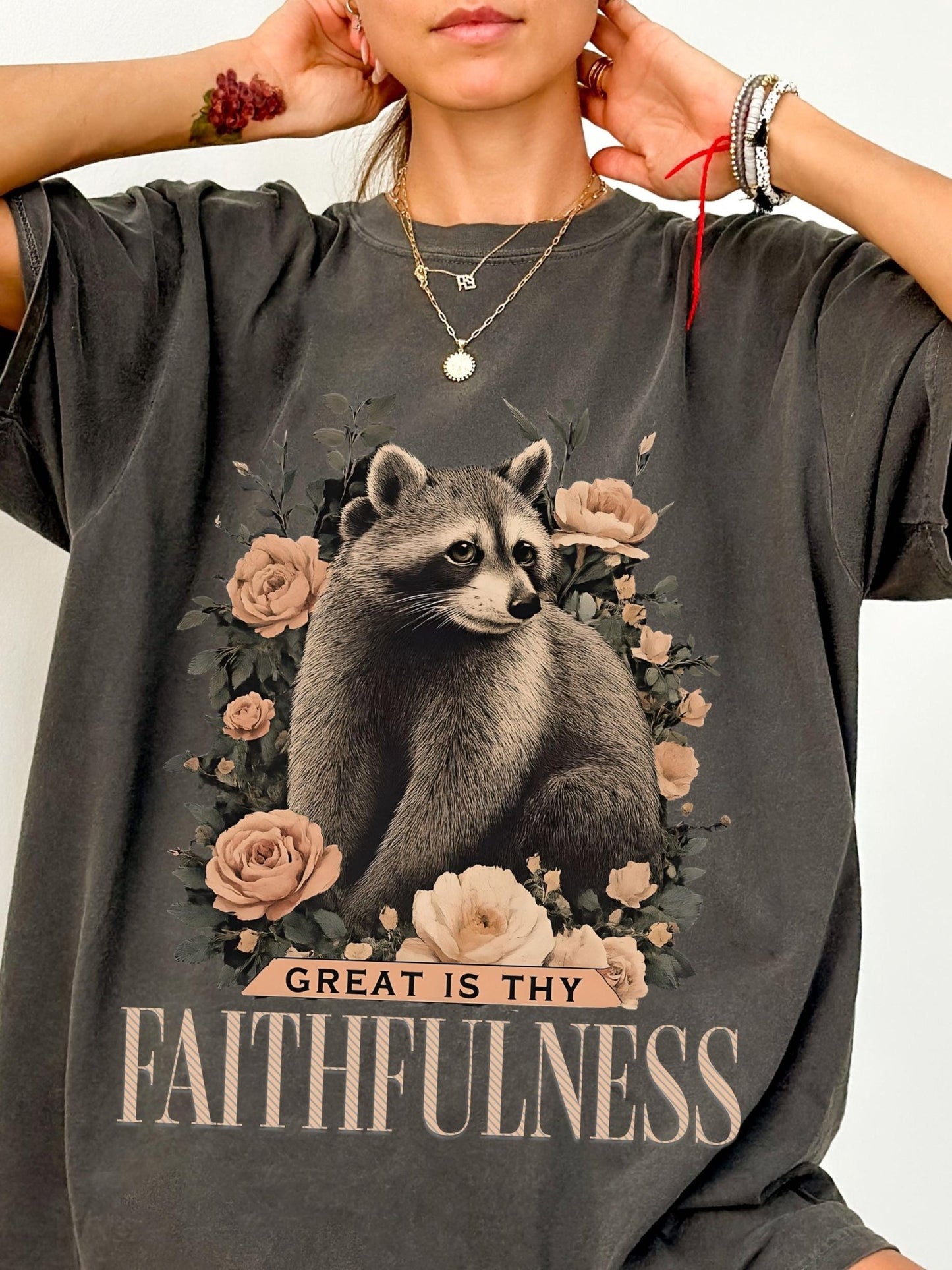 A person wearing a gray t-shirt with a graphic design of a raccoon surrounded by flowers and the text "GREAT IS THY FAITHFULNESS" printed on it.