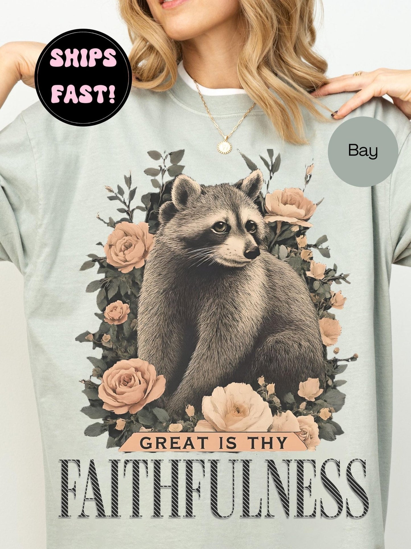 A person wearing a gray t-shirt with a graphic design of a raccoon surrounded by flowers and the text "GREAT IS THY FAITHFULNESS" printed on it.