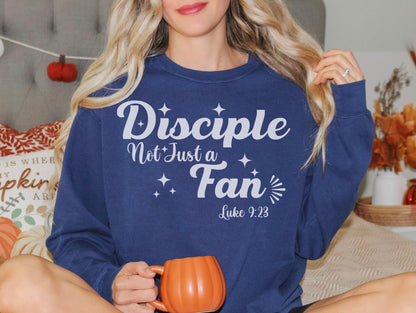 A woman wearing a blue sweatshirt with the text &quot;Disciples Not Just Fans&quot; and a Bible verse reference, &quot;Luke 9:23&quot;, is sitting on a bed with a pumpkin-themed pillow and holding a mug.