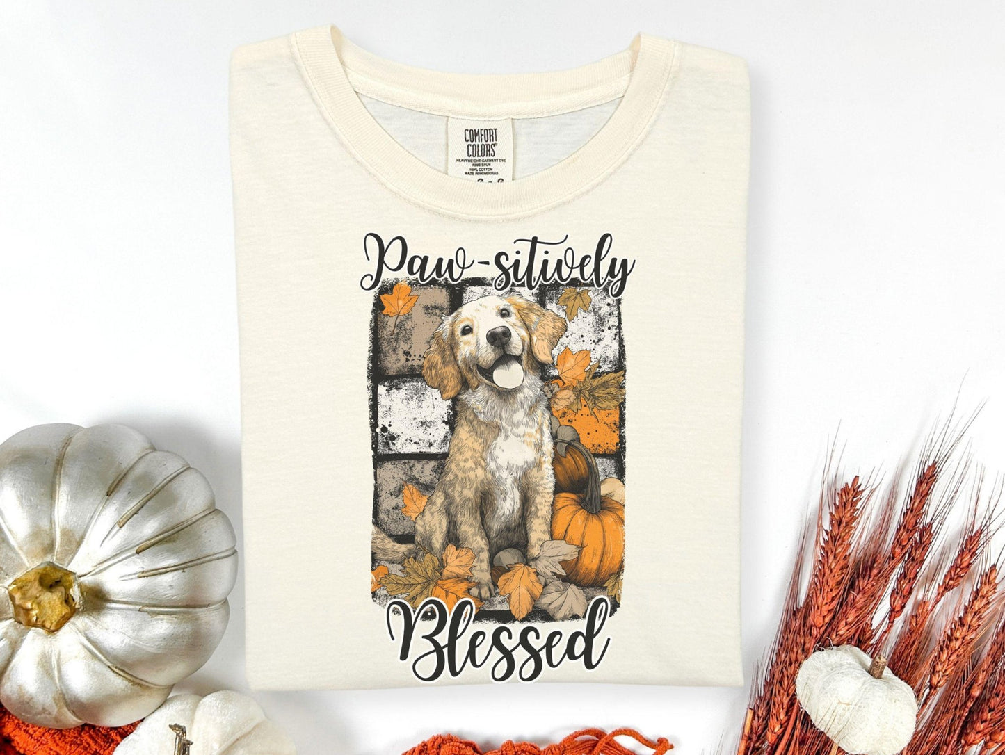 A white t-shirt with a graphic design featuring a dog surrounded by autumn leaves and pumpkins, along with the text "Paw sitively Blessed".