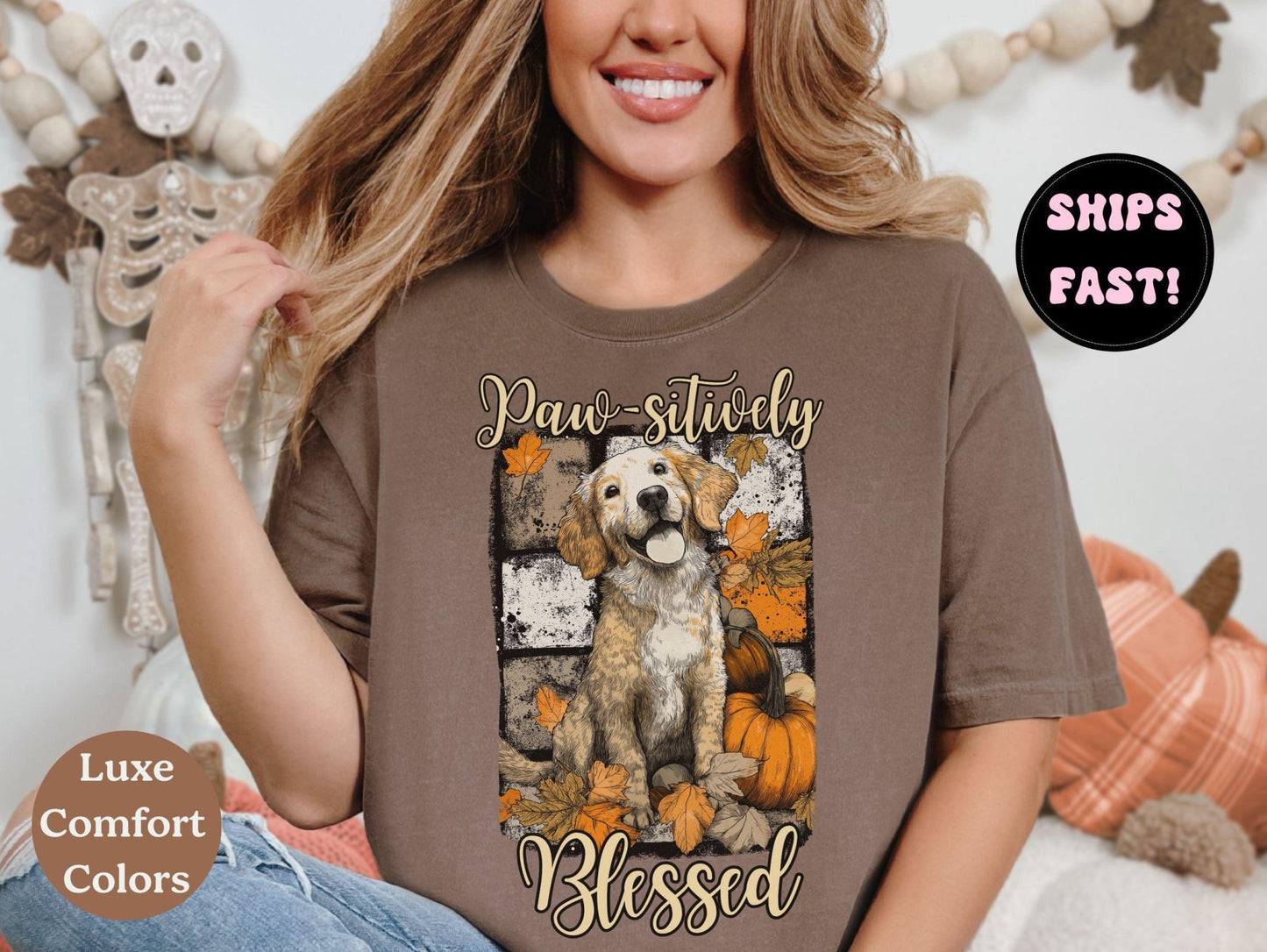 A woman wearing a brown t-shirt with a graphic of a dog surrounded by autumn leaves and pumpkins, with the text "Paw-sulively Blessed" printed on it.