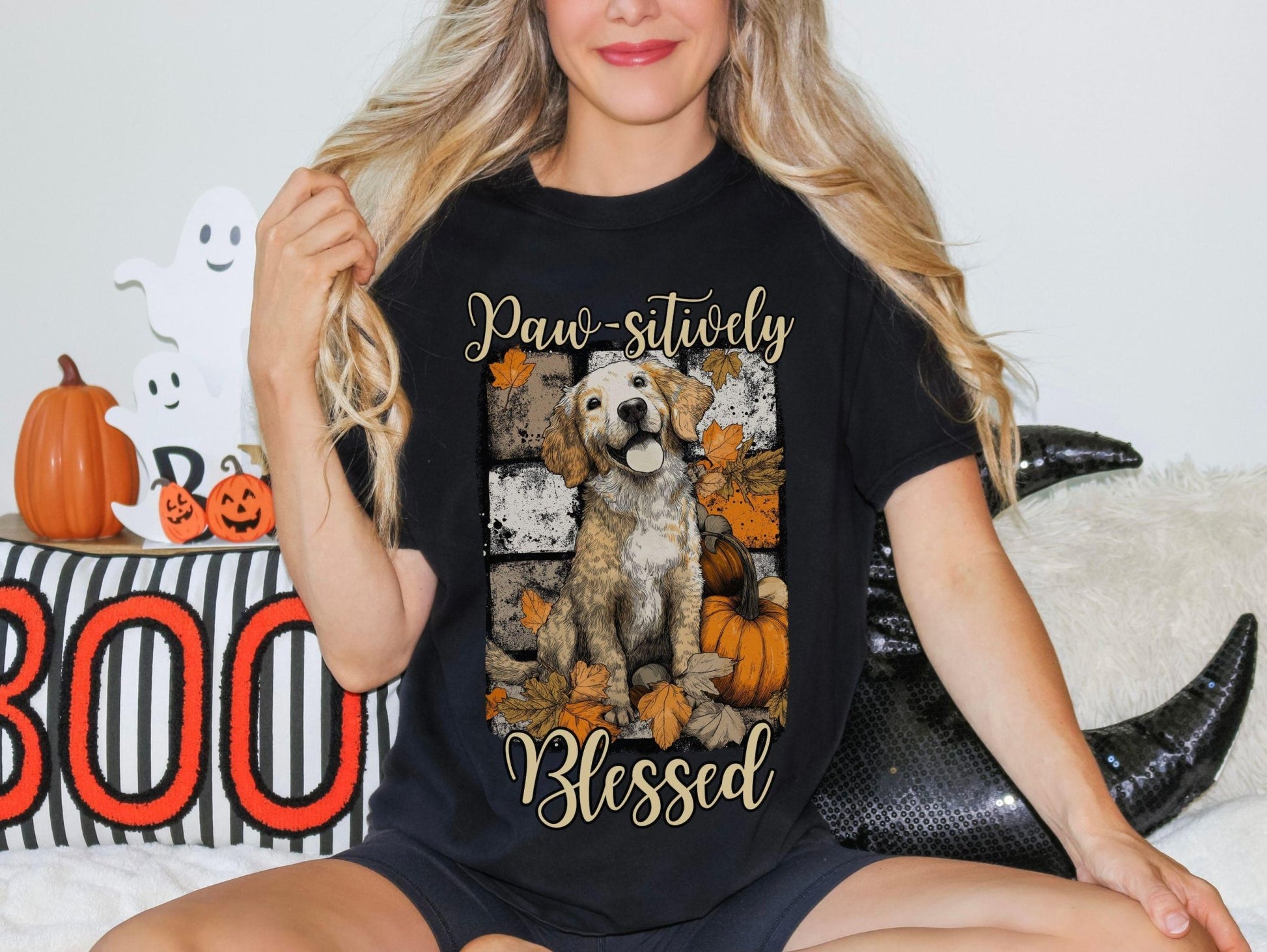 A woman wearing a black t-shirt with a graphic of a dog surrounded by autumn leaves and pumpkins, with the text "Paw-sitively Blessed" printed on it.