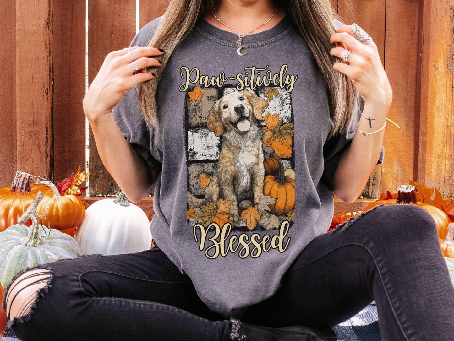 A woman wearing a gray t-shirt with a graphic of a dog surrounded by autumn leaves and pumpkins, with the text "Paw-sitively Blessed" printed on it.