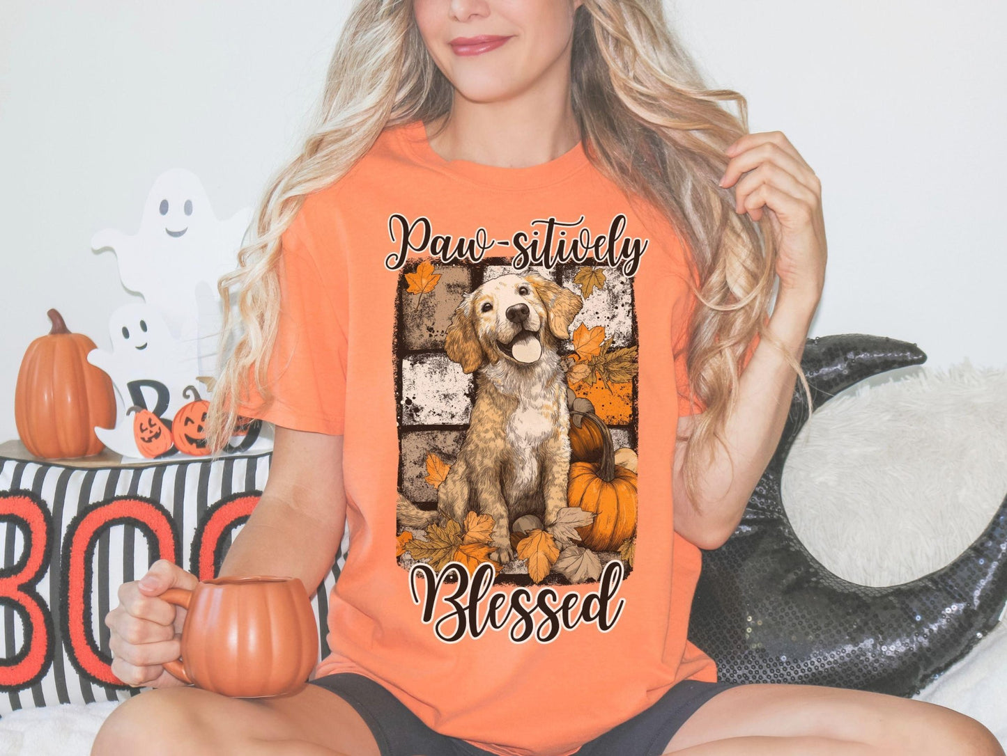 A woman wearing an orange t-shirt with a graphic of a dog surrounded by pumpkins and leaves, holding a pumpkin and a mug.
