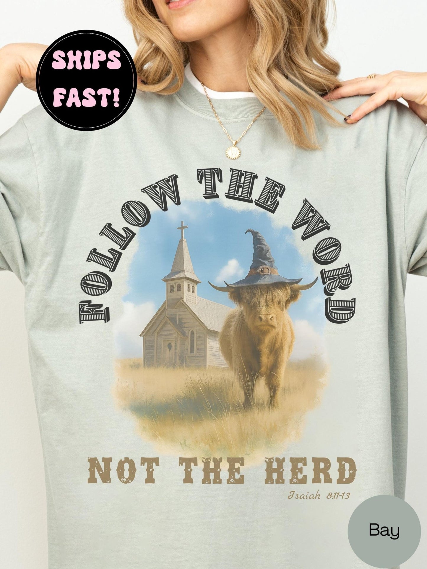 A woman wearing a bay green t-shirt with a graphic design featuring a cow wearing a witch&#39;s hat and a church in the background.