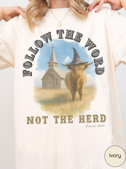 A person wearing a white t-shirt with a graphic design featuring a cow wearing a witch&#39;s hat and the text &quot;FOLLOW THE WORD NOT THE HERD&quot; along with a church in the background.