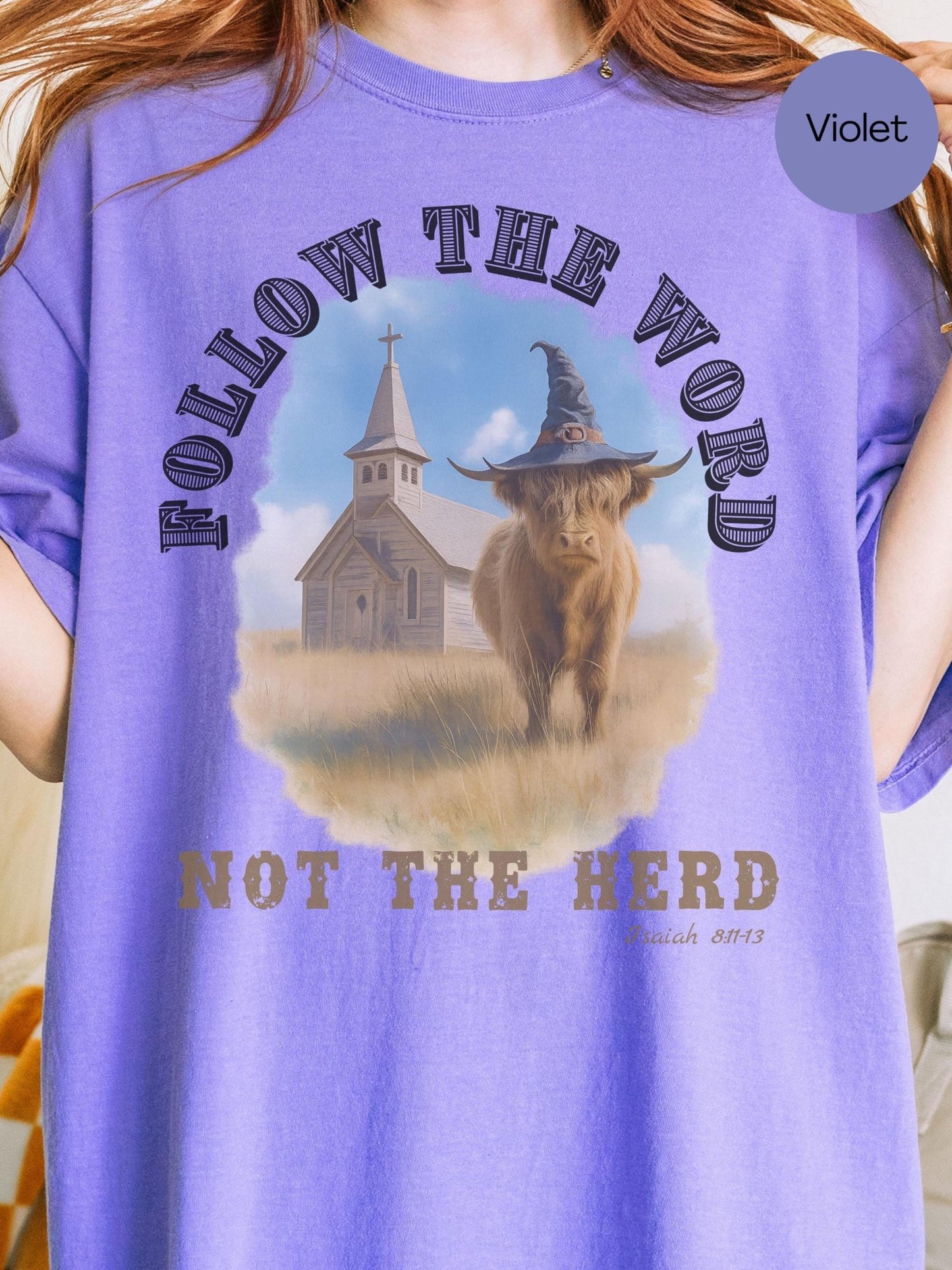 A purple t-shirt with a graphic design featuring a cow wearing a witch&#39;s hat and the text &quot;FOLLOW THE WORD NOT THE HERD&quot; along with a church in the background.