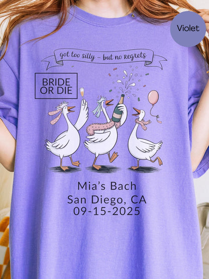 A purple t-shirt with a graphic design featuring three cartoon geese, one of which is holding a champagne bottle, surrounded by confetti. The text on the t-shirt reads &quot;got too silly, but no regrets&quot; and &quot;Bridal or Die&quot; along with &quot;Mia&#39;s Bach San Diego, CA 09-15-2025&quot;.