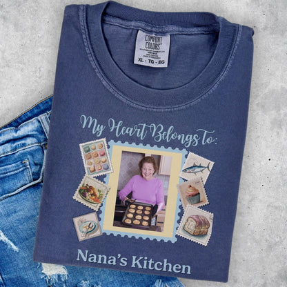 A blue t-shirt with a printed design that includes a photo of a woman, various food items, and postage stamps. The text on the t-shirt reads &quot;My Heart Belongs to: Nana&#39;s Kitchen&quot;.