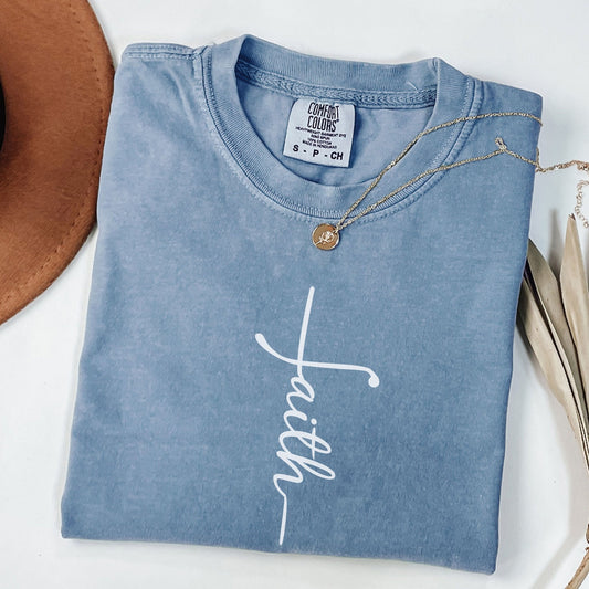 A blue t-shirt with the word "Faith" written in white cursive text is folded and displayed on a white surface, accompanied by a brown hat and a necklace.