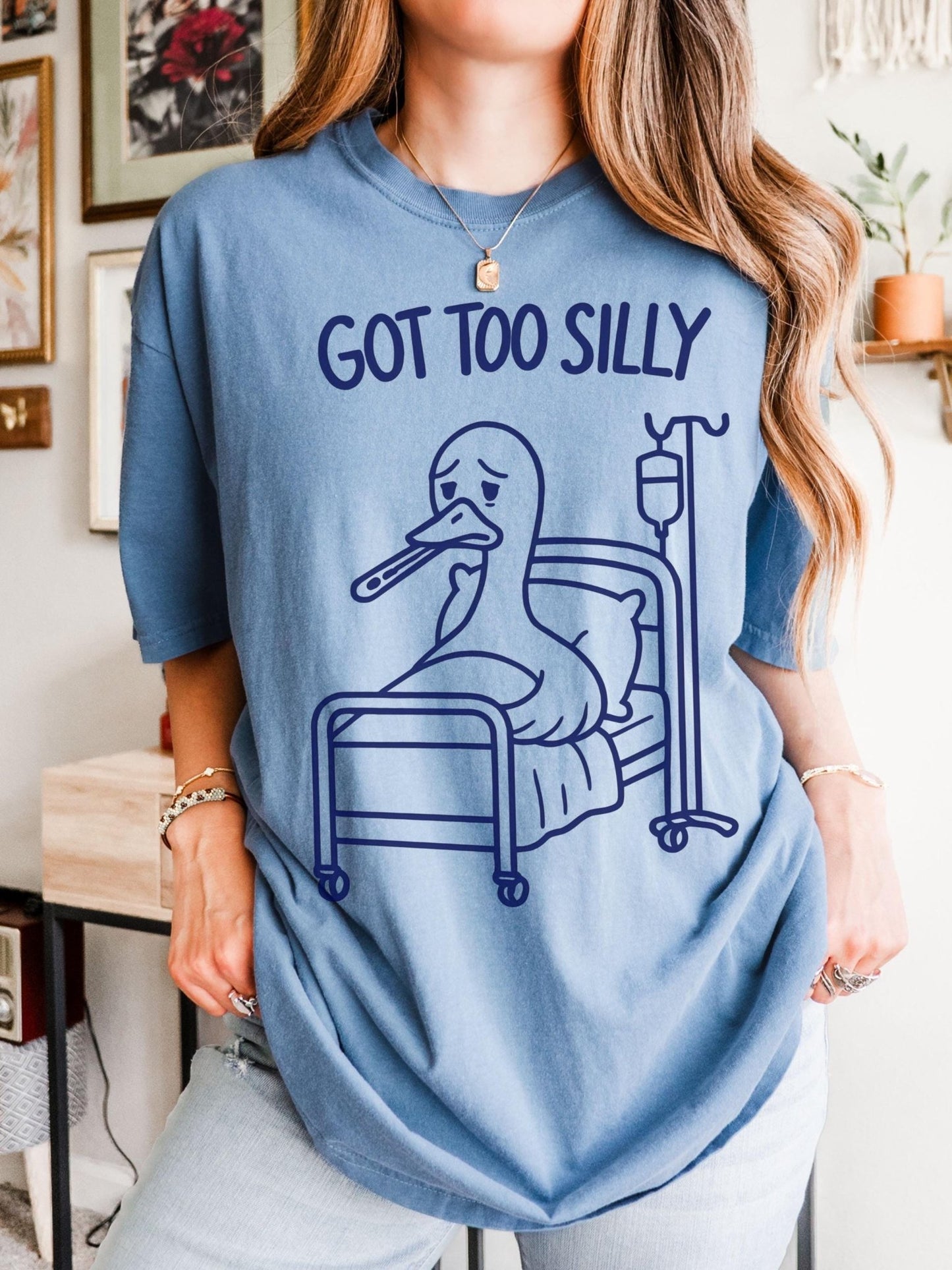 A woman wearing a blue t-shirt with a cartoon duck in a hospital bed and the text &quot;GOT TOO SILLY&quot; printed on it.