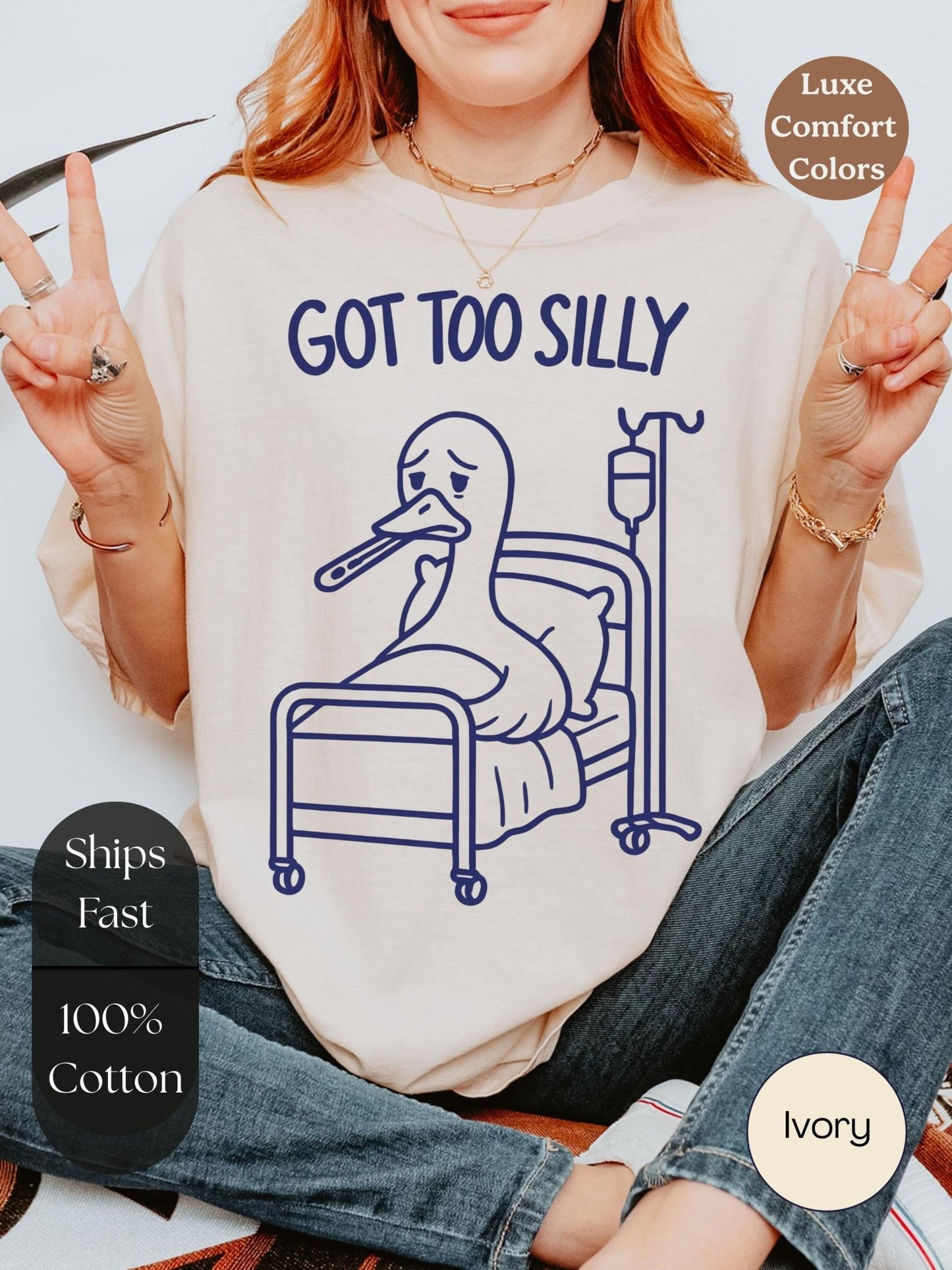 A woman wearing a white t-shirt with a graphic of a duck in a hospital bed and the text &quot;GOT TOO SILLY&quot; is making a peace sign.