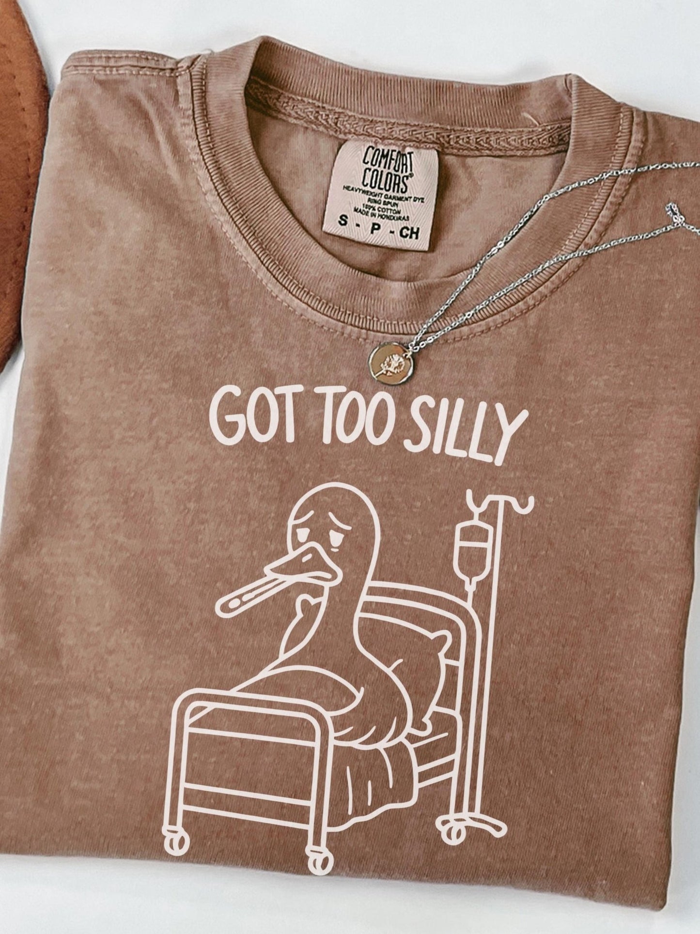 A brown t-shirt with a cartoon image of a duck in a hospital bed, accompanied by the text &quot;GOT TOO SILLY&quot; printed on it.