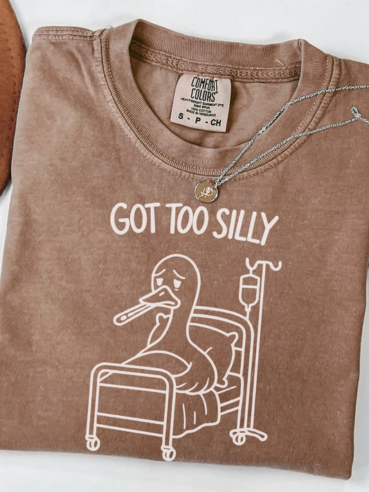 A brown t-shirt with a cartoon image of a duck in a hospital bed, accompanied by the text &quot;GOT TOO SILLY&quot; printed on it.