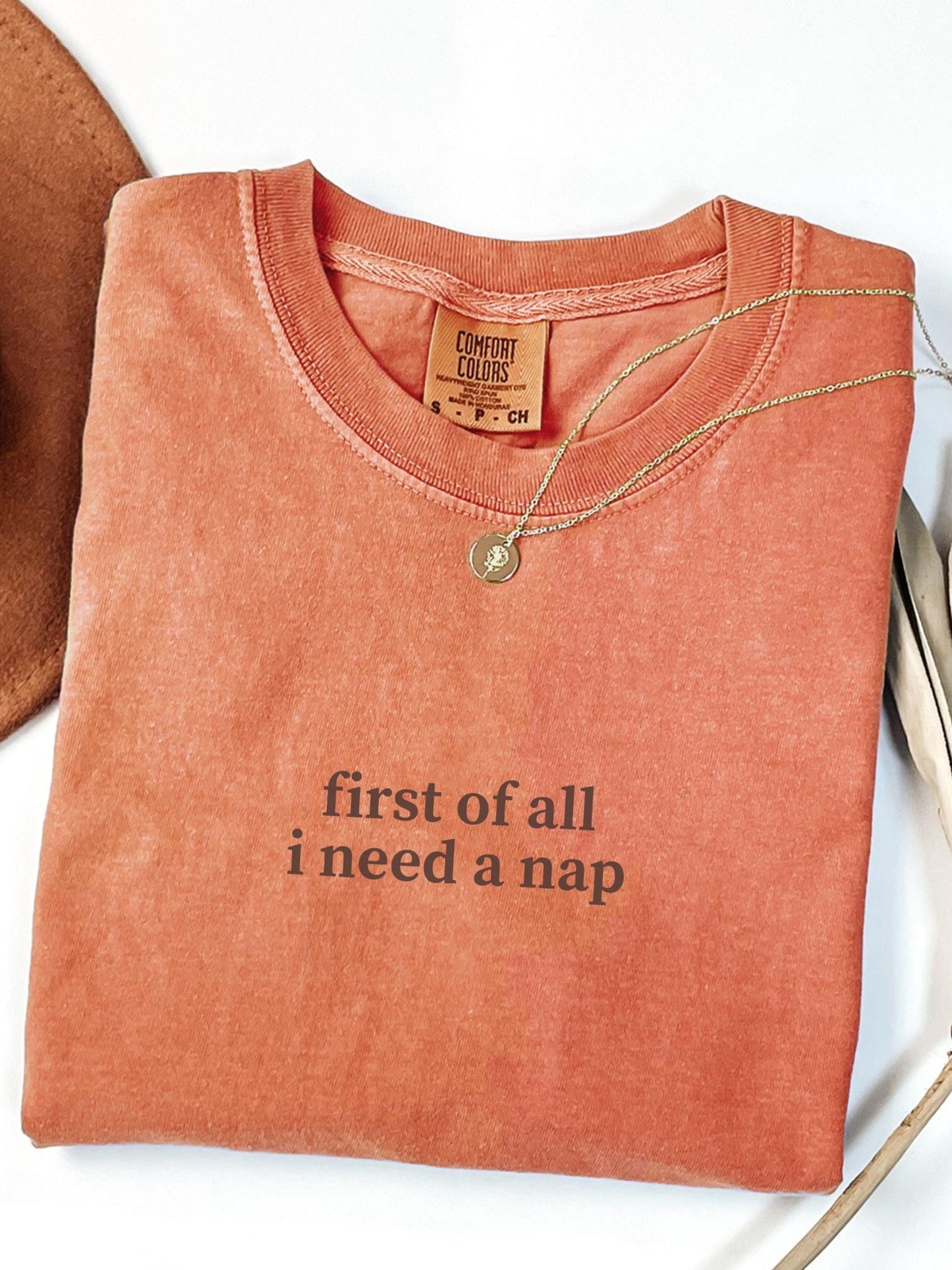 a red t-shirt with the text "first of all I need a nap" printed on it, along with a necklace and a hat.