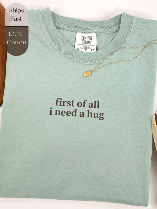 A light green t-shirt with the text &quot;first of all I need a hug&quot; printed on it, along with a gold necklace.