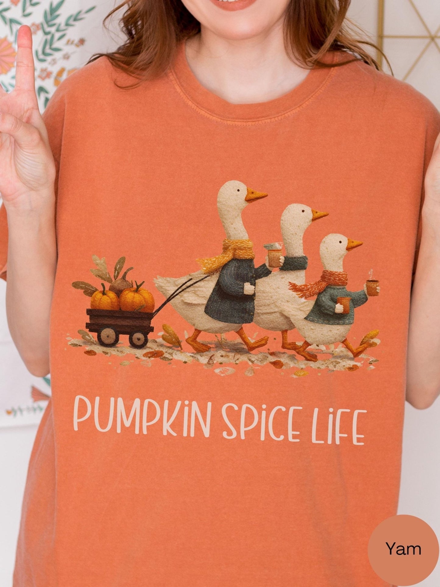 A woman wearing an orange t-shirt with a graphic design depicting three ducks walking on a path with a pumpkin carriage and holding pumpkins.