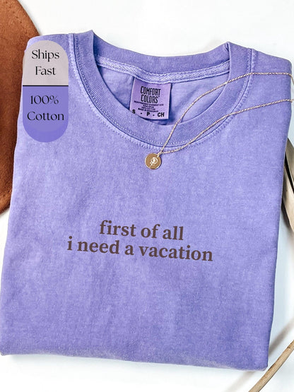 a purple t-shirt with the text "first of all I need a vacation" printed on it. The t-shirt is folded and displayed on a white surface, with a necklace visible around the neckline.