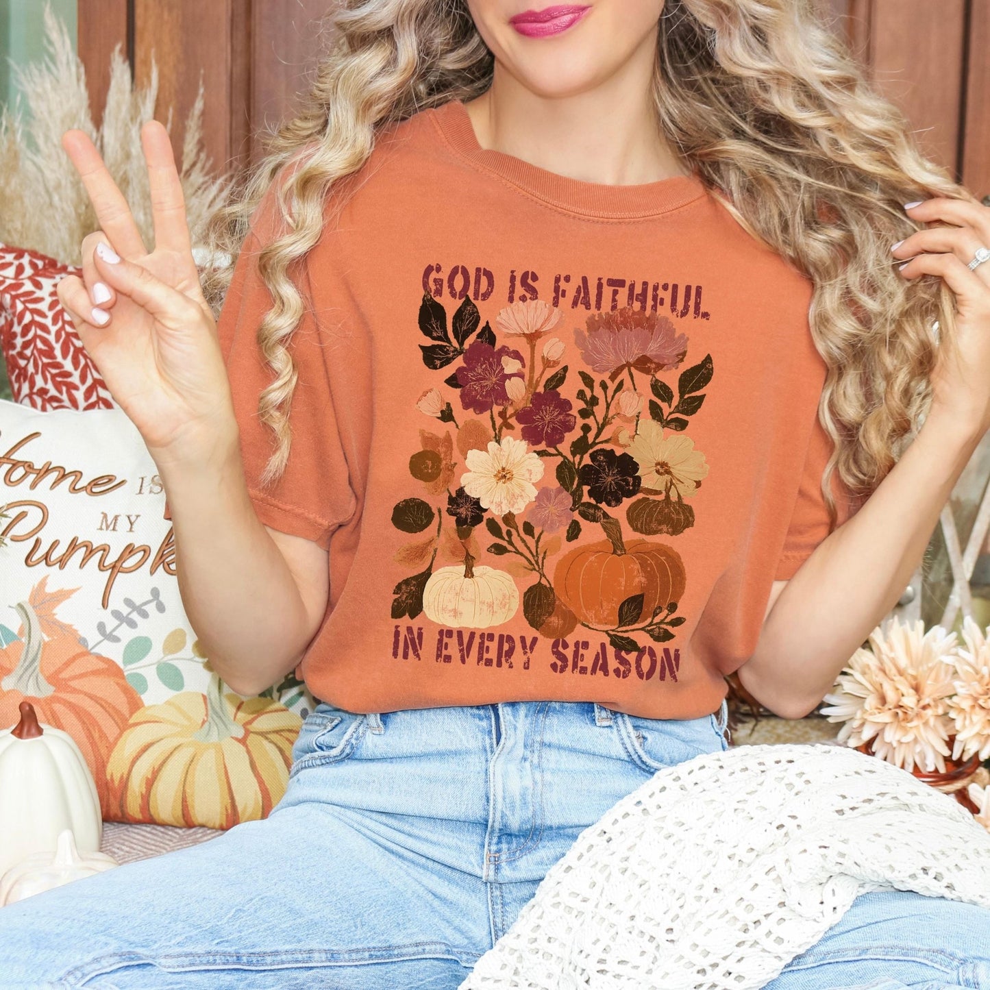 A woman wearing a t-shirt with a floral design and the text "God is Faithful in Every Season" is sitting on a couch surrounded by fall decorations.