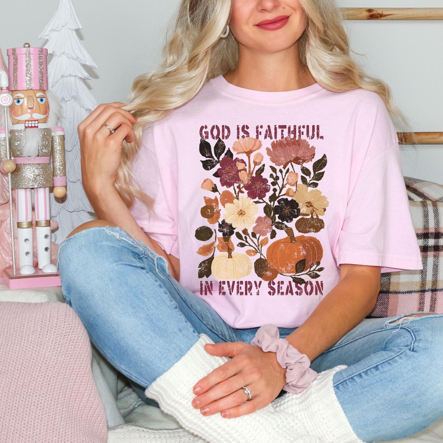 A woman wearing a pink t-shirt with a floral design and the text "God is Faithful in Every Season" is sitting on a couch.