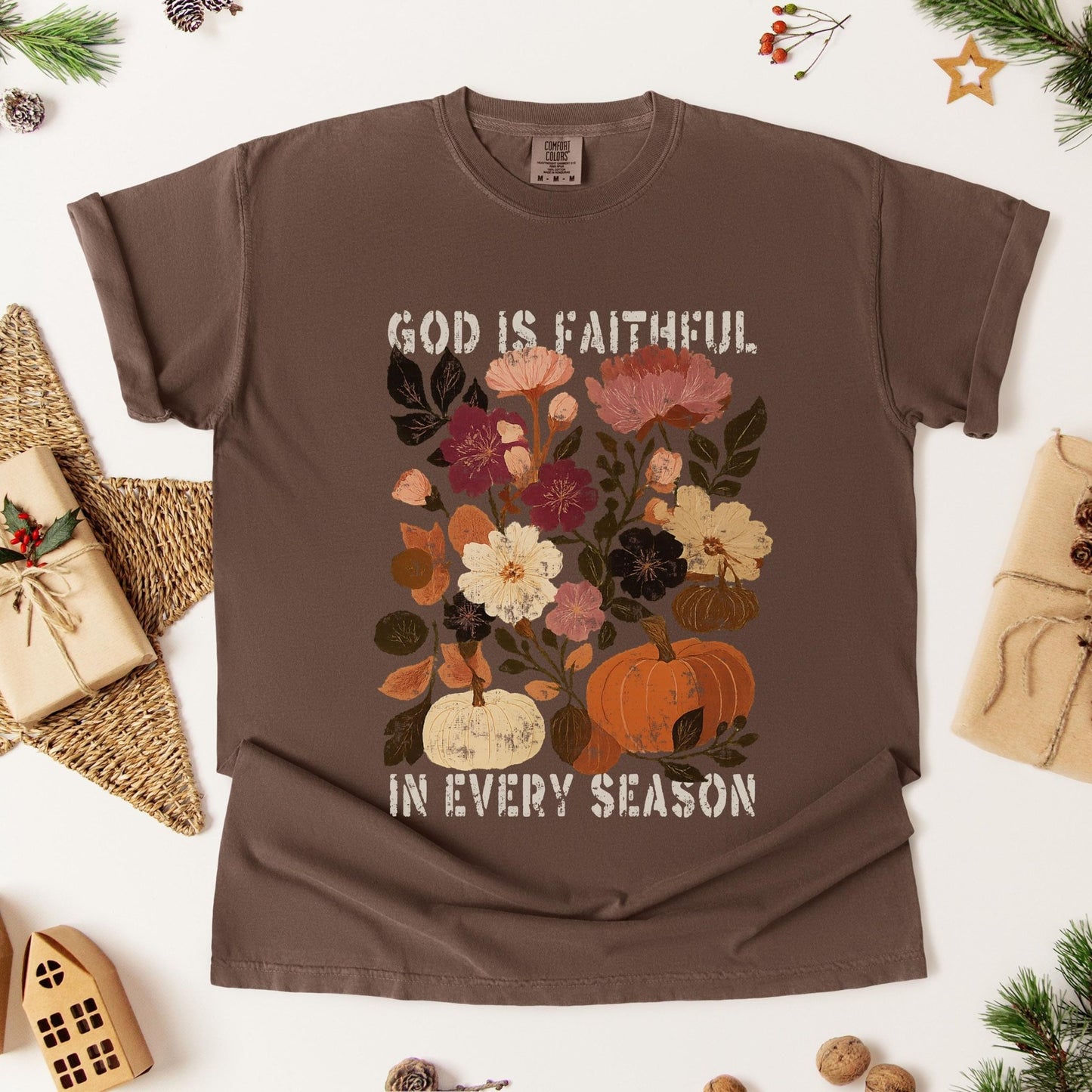 A brown t-shirt with a floral design and the text "GOD IS FAITHFUL IN EVERY SEASON" printed on it, along with a pumpkin and other fall-themed elements. The t-shirt is displayed on a white background, surrounded by various fall-themed items such as pine cones, a star, and a small house.