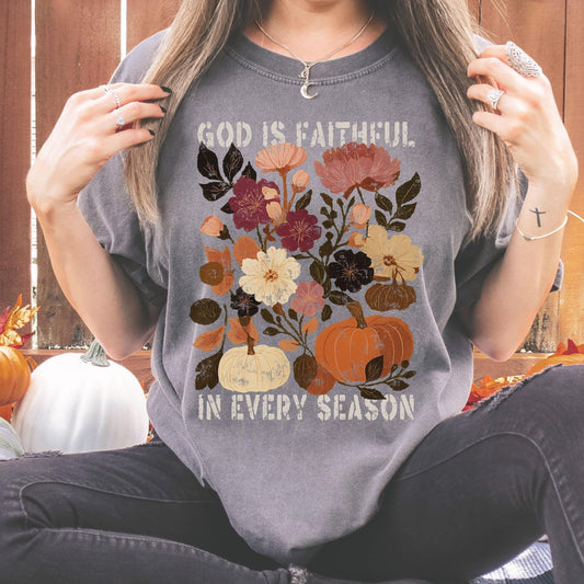 A woman wearing a gray t-shirt with a floral design and the text "God is faithful in every season" printed on it.