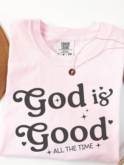 A pink t-shirt with the text "God is Good" printed on it, along with a small charm necklace.