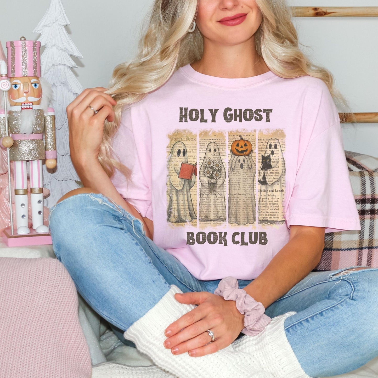 A woman wearing a pink t-shirt with a graphic of five ghostly figures and the text "HOLY GHOST BOOK CLUB" is sitting on a couch.
