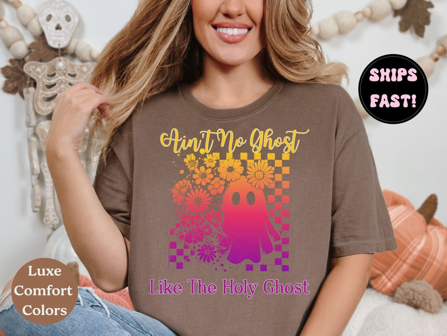 A woman wearing a brown t-shirt with a graphic design featuring a ghost and flowers, smiling and posing in front of a Halloween decoration.