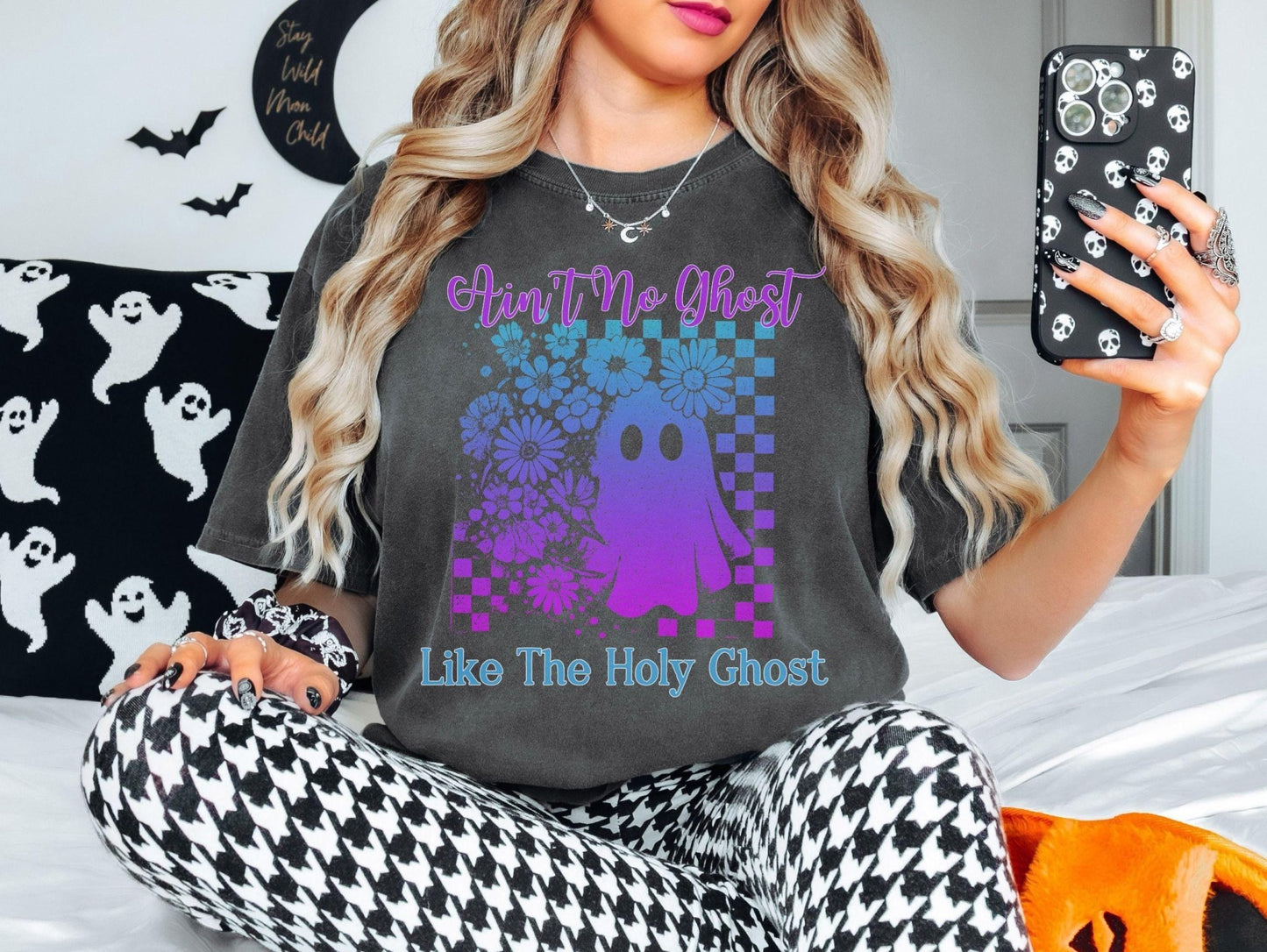A woman wearing a t-shirt with a graphic of a ghost and flowers, sitting on a bed and holding a phone.
