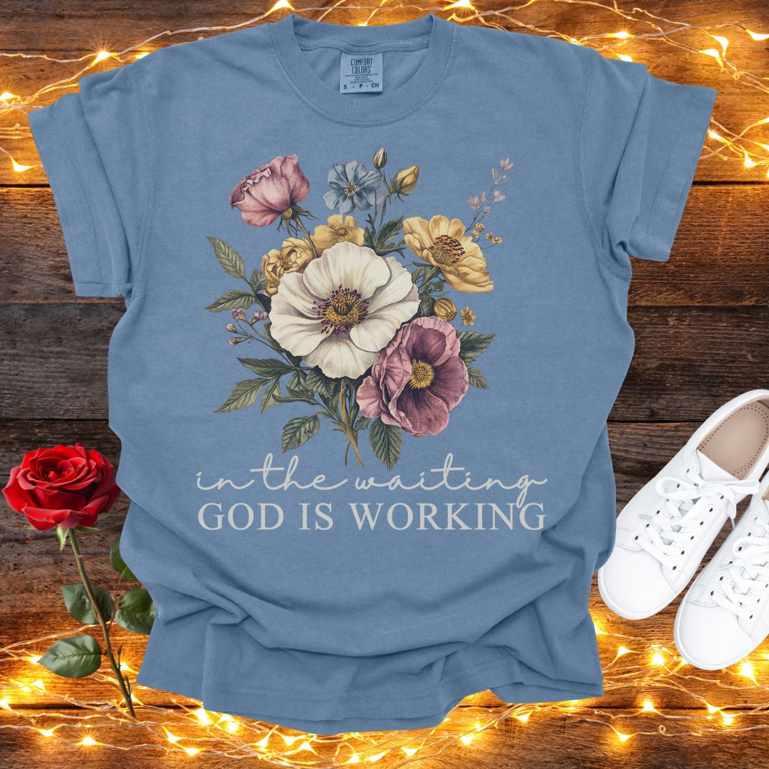 A blue t-shirt with a floral design and the text "in the waiting God is working" is displayed on a wooden surface, accompanied by a red rose and white sneakers.