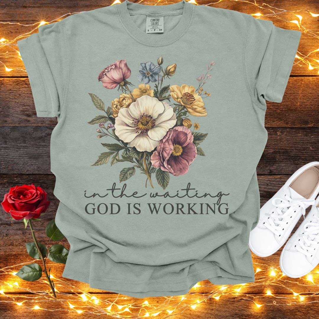 A green t-shirt with a floral design and the text "in the waiting God is working" is displayed on a wooden surface, accompanied by a red rose and white sneakers.