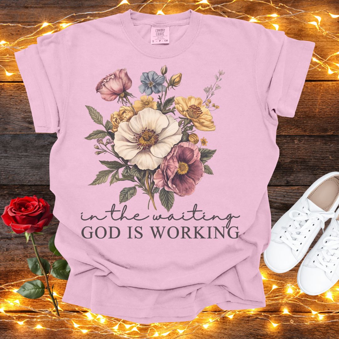 A pink t-shirt with a floral design and the text "in the waiting God is working" is displayed on a wooden surface, accompanied by a red rose and white sneakers.
