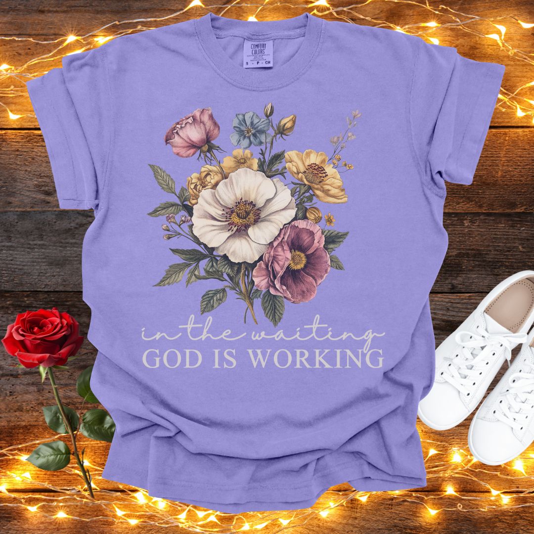 A purple t-shirt with a floral design and the text "in the waiting God is working" is displayed on a wooden surface, accompanied by a red rose and white sneakers.