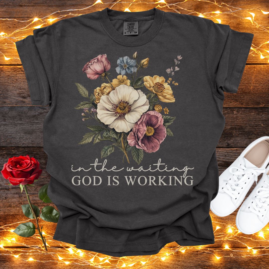 A black t-shirt with a floral design and the text "in the waiting God is working" printed on it, along with a pair of white sneakers and a red rose on a wooden surface.