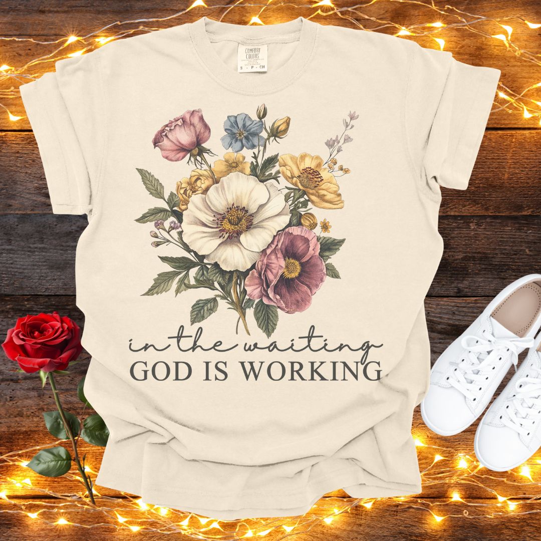 A beige t-shirt with a floral design and the text "in the waiting God is working" printed on it, along with a pair of white sneakers and a red rose on a wooden surface.